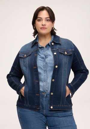 Frau trägt eine geschichtete Jeansjacke, Hemd und Jeans, steht mit den Händen in den Jackentaschen vor einem schlichten hellen Hintergrund.