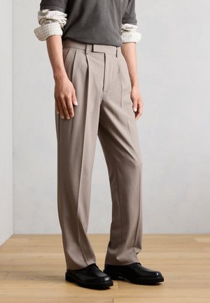 Man draagt beige geplooide broek, zwarte schoenen, donkergrijs overhemd over een wit geruit overhemd met lange mouwen, staat op een houten vloer.