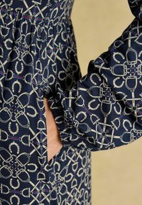 Marineblaues Kleid mit einem wiederholten Muster von Pferdegebissen und Zügeln, mit elastischen Ärmelbündchen und seitlichen Taschen. Baumwollstoff.