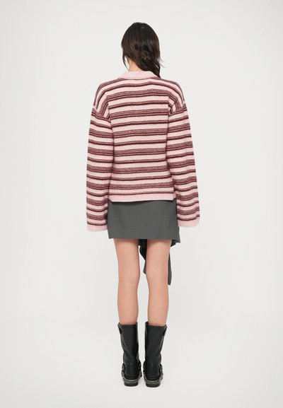 Ριγέ ζακέτα σε ροζ και μπορντό μαλλί με φαρδιά μανίκια, συνδυασμένη με γκρι φούστα και μαύρες μπότες μέχρι το γόνατο.