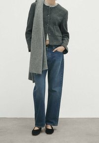 Veste grise texturée avec poches avant, associée à un haut gris clair, un jean bleu taille haute et des chaussures plates noires. Longue écharpe grise drapée par-dessus.