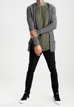 Mann mit tätowiertem Hals und Händen, trägt ein grünes Hemd, eine graue offene Strickjacke, schwarze Hose und schwarz-weiße Sneakers, steht mit gespreizten Beinen.