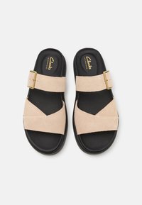 Slide sandals with a tan suede upper featuring a crisscross design. Black rubber sole, gold-tone buckle accent, and soft textured footbed.