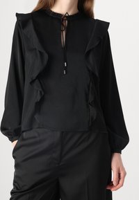 Blouse noire avec manches à volants, col rond et une ouverture en forme de trou de serrure à l'avant. Dispose d'une fermeture à cordon avec des embouts métalliques. Tissu lisse.