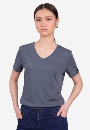 Giovane donna con capelli lisci e scuri che indossa orecchini a cerchio dorati, una maglietta grigia con scollo a V e jeans blu scuro con le mani nelle tasche.
