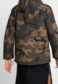 Camouflage capuchonjack in tinten van groen, bruin en zwart, met een ritssluiting en een koordverstelling aan de onderkant.