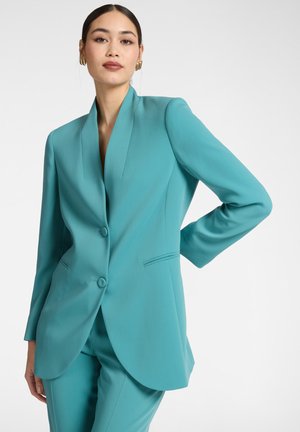 Frau in einem türkisfarbenen, maßgeschneiderten Anzug mit zweiknöpfigem Blazer und passenden Hosen, die mit einer Hand in der Hüfte vor einem schlichten Hintergrund posiert.