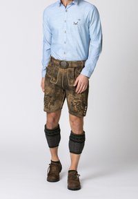 Muž oblečený v tradičních hnědých kožených lederhosen s výšivkou na kšandách, světlomodré košili s vzorem, pletených zateplovačích na lýtka a hnědých botách.