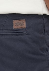 Gros plan sur la poche d'un pantalon bleu marine avec une étiquette marron de la marque Jack & Jones cousue au-dessus de la couture de la poche et une partie d'une chemise blanche visible.