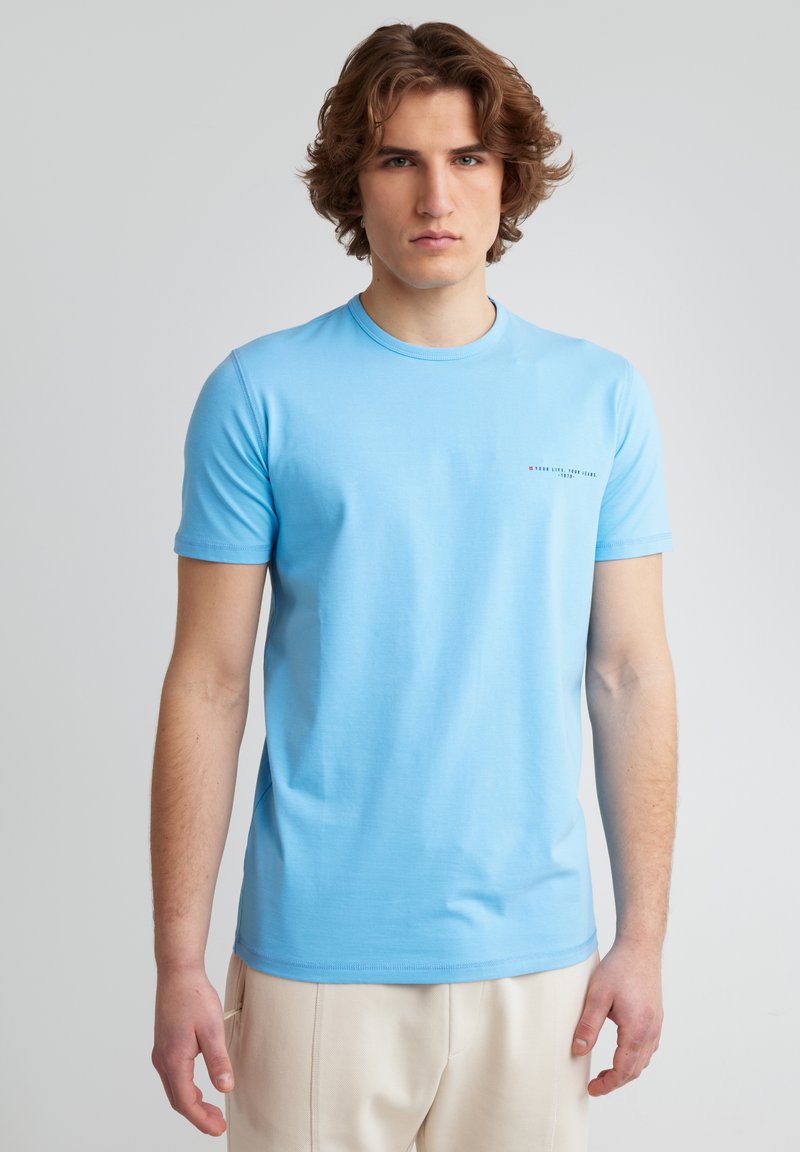 Gaiši zils īsām piedurknēm t-krekls, kas izgatavots no kokvilnas. Tam ir apaļa kakla izgriezums un neliels drukāts logo kreisajā krūšu pusē.