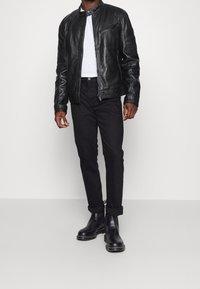 Uomo con giacca di pelle nera, t-shirt bianca, pantaloni neri arrotolati e stivali di pelle nera, in piedi contro uno sfondo chiaro e semplice.