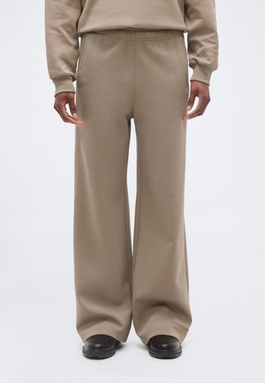 Isik, kes kannab lahtisi beeže püksklambreid elastse vöökohaga ja musti jalatseid, seisab ühevärvilise valge tausta ees.