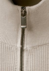 Detailaufnahme eines beigen, gerippten Strickpullovers mit einem teilweise geschlossenen Reißverschluss aus silberfarbenem Metall.