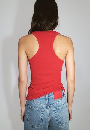 Femme aux longs cheveux foncés portant un débardeur rouge dos nageur et un jean taille haute bleu clair, debout devant un fond gris uni.