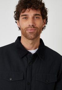 Zwarte jas met een corduroy kraag, voorzien van twee borstzakken en drukknopen, gemaakt van een gladde stof met een schoon ontwerp.