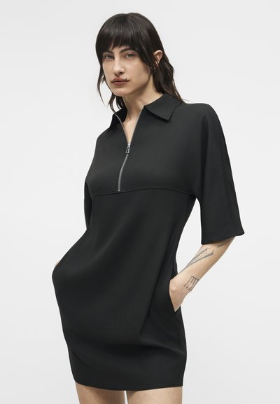 Νεαρό άτομο με σκούρα μαλλιά που φοράει μαύρο φόρεμα με κοντά μανίκια και φερμουάρ, με τα χέρια στις τσέπες, πάνω σε απλό φωτεινό φόντο.