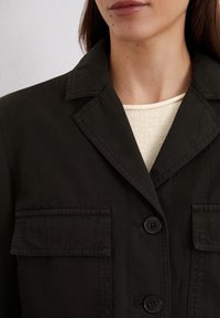 Svart bomullsjacka med krage, två framfickor och knäppning med knappar, bärs över en ljus stickad topp. Mjuk textur.