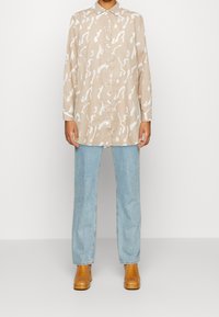 Chemise beige à manches longues avec un motif abstrait blanc, devant à boutons et col classique, associée à un jean bleu clair et des bottines beiges.