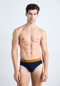 Marineblauwe slips met een beige elastische tailleband met branding. De stof lijkt soepel met een getailleerd ontwerp. Het model heeft een slank figuur.