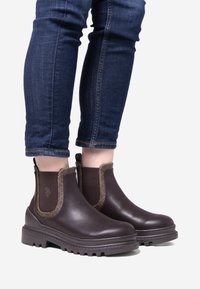 Hnědé kožené Chelsea boty s elastickými bočními panely a texturovaným lemem. Mohutná gumová podrážka, nošeny s tmavě modrými džíny.