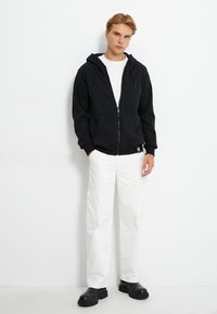 Sudadera negra con cremallera y capucha, bolsillos frontales y puños de canalé, combinada con pantalones cargo blancos y botas negras de suela gruesa.