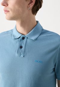 Tânăr îmbrăcat cu un tricou polo albastru deschis, cu două nasturi încheiați și logo-ul "BOSS" pe piept, întors spre stânga, purtând un cercei în formă de cerc.