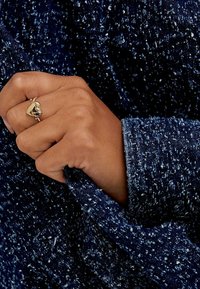 Tessuto navy con una trama texturizzata e un motivo a macchie. Una mano afferra la manica, mostrando un anello d'oro con un design a forma di cuore e una pietra scura.