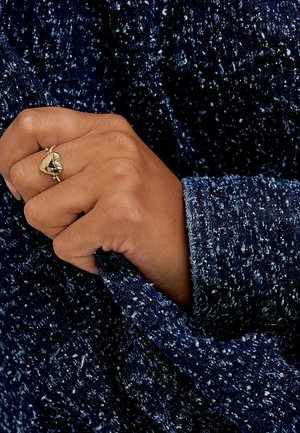 Marinblått textil med en spräcklig mönstring. En hand greppar ärmen och visar en guldring med ett hjärtformat mönster och en mörk ädelsten.