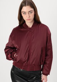 Giacca bomber bordeaux con una texture liscia e lucida, colletto e polsini a costine, zip frontale e dettagli con tasche laterali.