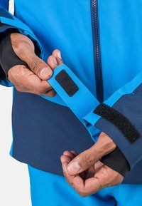 Blau wasserdichte Jacke mit einem Reißverschluss vorne und verstellbaren Manschetten mit Klettverschluss, kombiniert mit einem dunkelblauen unteren Abschnitt.