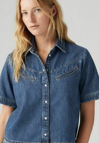 Camicia di jeans a maniche corte, blu con una finitura texture. Presenta una chiusura con bottoni davanti, due tasche sul petto e una cucitura a contrasto lungo le cuciture.
