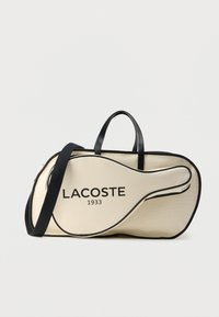 Beżowa torba w kształcie rakiety tenisowej z czarnymi akcentami i logo "LACOSTE 1933", posiadająca podwójne uchwyty oraz pasek na ramię do przenoszenia.