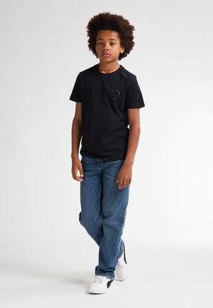 T-shirt en coton noir avec un logo rond sur la poitrine, associé à un jean bleu et des baskets blanches. Enfant se tenant devant un fond clair.