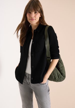 Kobieta ubrana w czarną, zapinaną na guziki kurtkę na białą bluzkę, szare dżinsy, niosąca zieloną pikowaną torebkę na ramię, na beżowym tle.