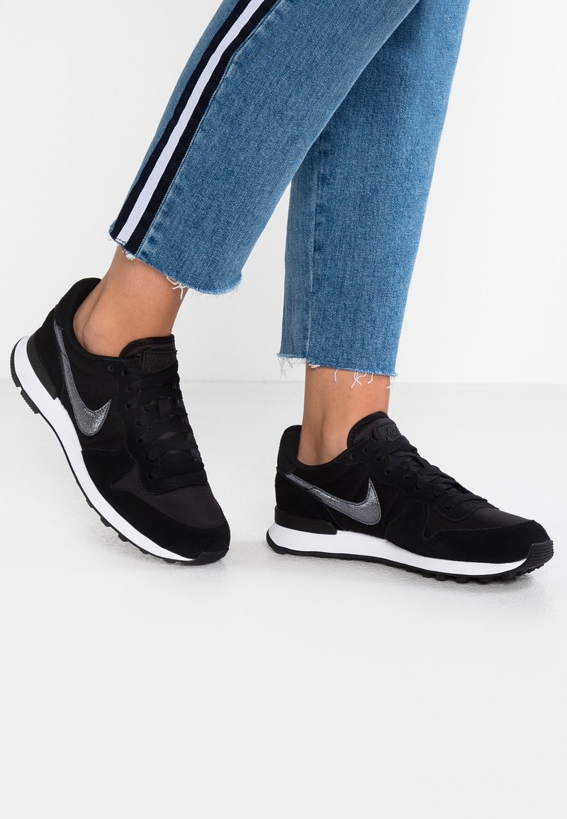 Personne portant un jean bleu avec des bandes latérales noires et blanches et des baskets Nike noires avec des semelles blanches sur un sol blanc.
