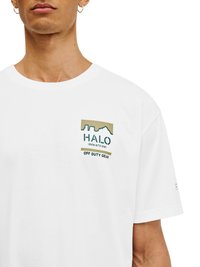 Mężczyzna w białej koszulce z napisem "HALO 198819721981 OFF DUTY GEAR" i grafiką miasta na klatce piersiowej.