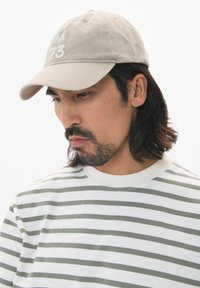 Căciulă de baseball bej cu o șapcă curbată și un design brodat. Modelul poartă o cămașă albă cu dungi verzi.