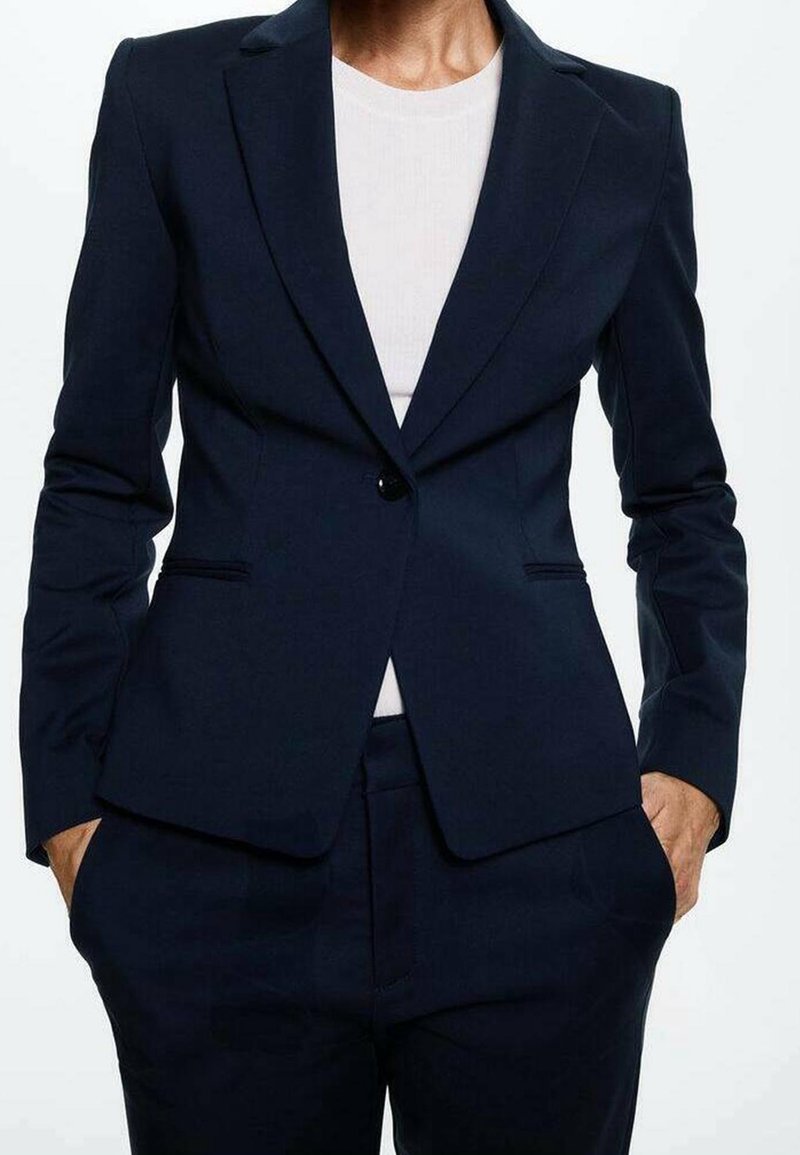 Personne portant un blazer ajusté bleu marine foncé sur une chemise blanche, les mains dans les poches du pantalon bleu marine assorti.