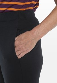 Pantaloni neri con una texture liscia e vita elastica. Mano posizionata in una tasca laterale, evidenziando un design slanciato.