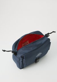 Marineblaue Stofftasche mit Reißverschluss, rotem Innenfutter, verstellbarem Trageriemen und Logo-Detail auf der Vorderseite. Kompakte rechteckige Form.
