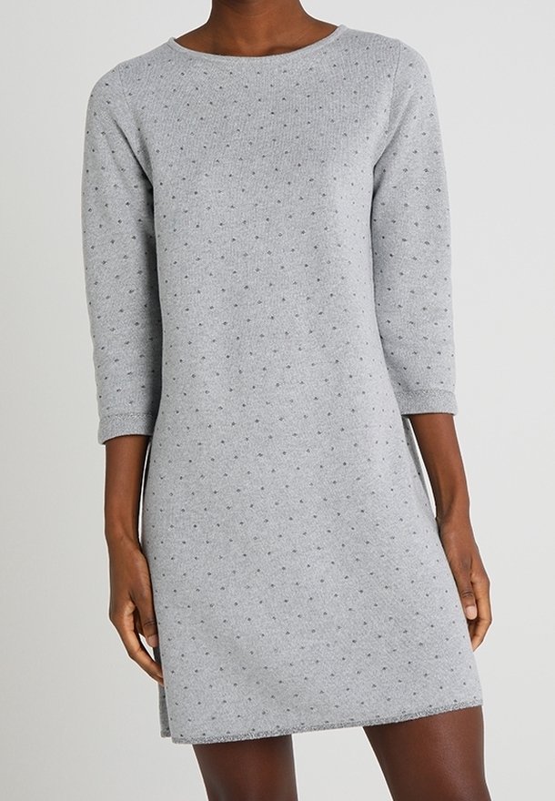 Femme portant une robe grise, à manches longues, longueur genoux, avec un motif de petits points subtilement perceptible, les mains reposant le long du corps.