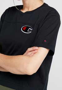 Černé tričko s krátkým rukávem vyrobené z bavlny s kulatým výstřihem, s výrazným vyšitým logem v červené a bílé barvě na hrudi.