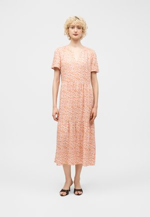 Kobieta o kręconych blond włosach, ubrana w różowo-pomarańczową sukienkę midi w wzory oraz czarne, odkryte buty na obcasie, stojąca na jasnym, jednolitym tle.