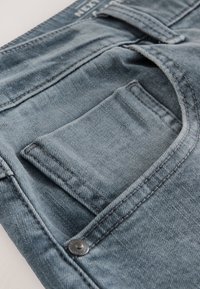 Blaue Jeans aus Denim mit sichtbarer Gesäßtasche und Nahtdetails. Verfügt über einen Metallknopf und strukturierten Stoff. Draufsicht liegt auf der Tasche.