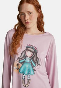 Roodharige vrouw met sproeten draagt een lichtroze shirt met lange mouwen, waarop een cartoonmeisje te zien is in een teal jurk dat een lantaarn vasthoudt met een vlinder erbij.