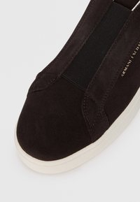 Black suede sneaker with elastic side panel and white rubber sole. Features stitching detail and logo in gold on the side.
