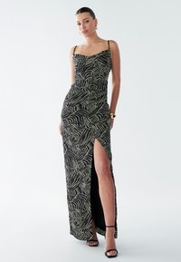 Black maxi dress with a leaf pattern in cream, featuring a fitted design, spaghetti straps, and a thigh-high slit. Smooth, lightweight fabric.