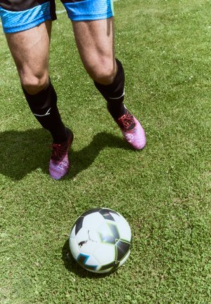 Jugador de fútbol con pantalones cortos negros con acentos azules, usando zapatillas multicolores, pateando un balón de fútbol con un patrón en blanco y negro sobre hierba.