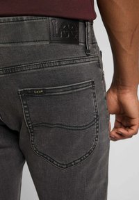 Jeans in denim grigio scuro con una patch in pelle del marchio Lee sulla vita. Presentano due tasche posteriori con dettagli di cucitura visibili.