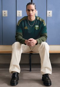 Groene sporttrui met gouden strepen en logo, crèmekleurige wijde broek en zwarte veterschoenen. Geplaatst tegen kluisjes met nummers.